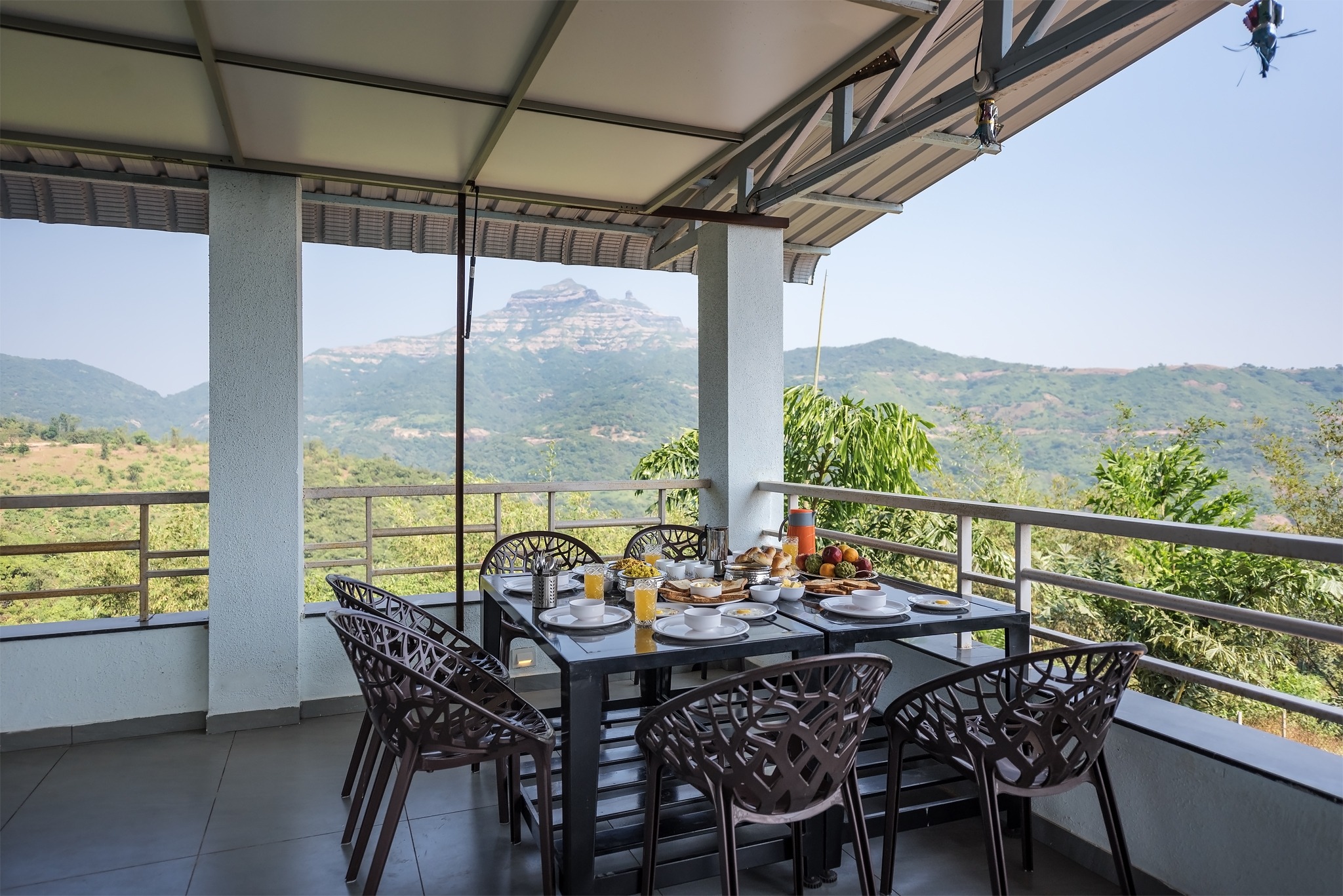 A covered outdoor dining area with a table set for breakfast, surrounded by chairs. The space overlooks green hills and a distant mountain under a clear blue sky.