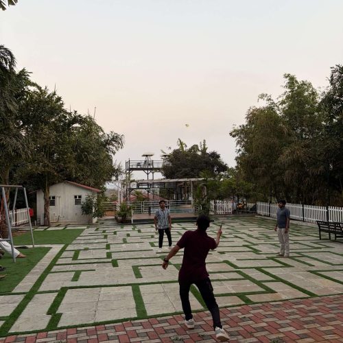 Four people are playing badminton outdoors on a tiled patio surrounded by trees and a white fence, with a shed and scaffolding in the background under a clear sky.