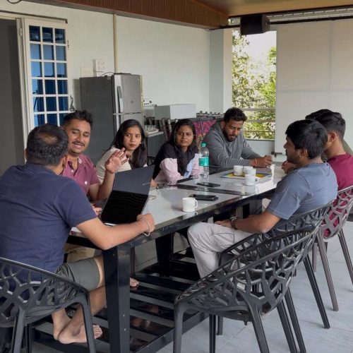 A group of eight people sit around a rectangular table on a covered patio, engaged in conversation and smiling. Laptops, notebooks, cups, and a water bottle are on the table. Natural light fills the modern space.