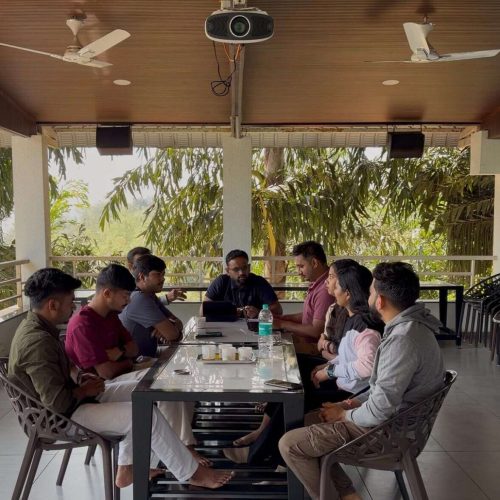 A group of nine people sit around a rectangular table on a covered patio, engaged in conversation. Trees are visible outside. The time, date, and location are displayed at the top of the image.