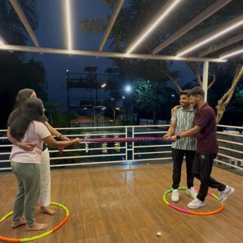 Four people stand in pairs inside two separate hula hoops on a wooden deck at night, with string lights overhead and trees visible in the background. Each pair faces each other, appearing engaged in an activity.