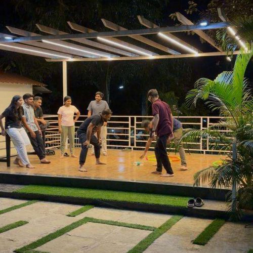 A group of people are playing an indoor game with a stick and ball on a wooden deck at night, while others watch. The area is lit with overhead lights and surrounded by greenery.