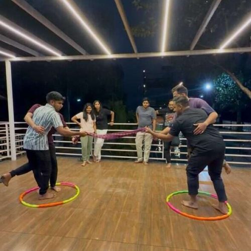 A group of people play a game with colorful hula hoops on a wooden floor under outdoor lights at night, while onlookers watch and smile in the background.